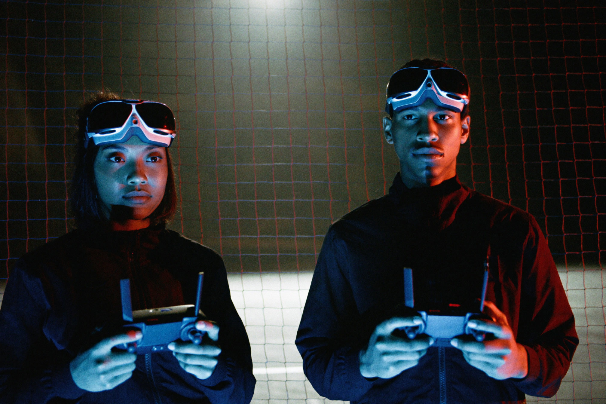 Two pilots in FPV goggles holding controllers outside the arena net